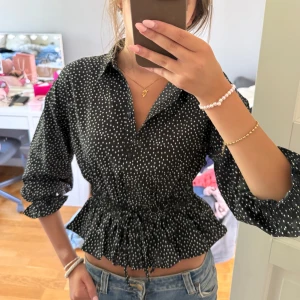Svart prickig blus från Pigalle XS - Supersöt svart blus från Pigalle med vita prickar, markerad midja med snörning och volangkant nertill. 