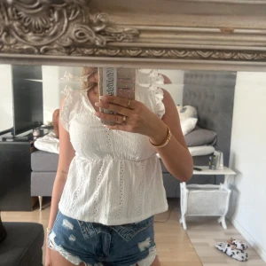 Vit blus med volangärmar - Supersöt vit blus med v-ringning och volangärmar. Toppen har broderade detaljer och lätt rynkad midja som ger en feminin siluett. Perfekt för varma dagar och ger en romantisk vibe till din outfit.