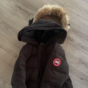 Svart dunjacka från Canada Goose - Säljer en svart dunjacka från Canada Goose med klassisk patch på ärmen och stor huva med fluffig pälsimitation. Jackan är lång, har dragkedja och knappar framtill och är perfekt för kalla vinterdagar. Snygg och tidlös modell som håller dig varm hela säsongen. Lite skada på armen men de inget märkvärdigt. Jackan är äkta självklart priset går att diskutera vid frågor hör av dig:)