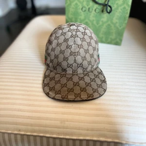 Gucci keps  - Exklusiv keps från Gucci i beige och brunt med klassiskt GG-monogram över hela kepsen. Kepsen har gröna och röda ränder på sidan och är tillverkad i bomullsmix. Perfekt för dig som vill sticka ut med en ikonisk accessoar. Varan är så klart äkta! Finns lite defekter på kepsen priset är inte hugget i sten!