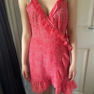 Röd omlottklänning med volang - Söt röd kort omlottklänning med små vita blommor, volangkant nertill och knyt i sidan. Skriv vid intresse!