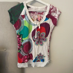 Färgglad blommig topp från Desigual - Unik och färgstark topp från Desigual med blommigt och grafiskt mönster i grönt, rött, lila, blått och vitt. Rund halsringning med knyt och korta ärmar. Materialet är mjuk bomull, perfekt för dig som gillar statement-plagg och kreativa prints. Passar till yk2 style och nypris är 200kr, pris går att diskutera!