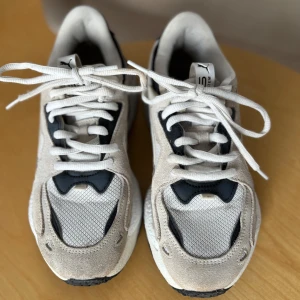 Puma vita och svarta sneakers - Chunky sneakers från Puma i vitt och svart med snygga detaljer i mocka och mesh. Skorna har klassisk snörning och en robust sula som ger en cool streetstyle-vibe. Perfekta för dig som gillar sportig och trendig look.