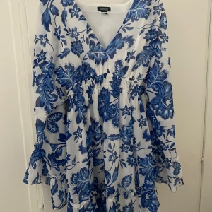 Blåvit blommig kortklänning Boohoo - Supersöt kortklänning från Boohoo med blått blommönster på vit botten. Klänningen har v-ringning, långa vida ärmar med volang och smock i midjan för en snygg passform. Perfekt för sommarens alla tillfällen!