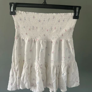 Två volangkjolar med smock i midjan - Säljer två söta korta kjolar med smock i midjan och volanger. En är vit med små blommor i rosa och blått, den andra är helsvart. Båda är i mjukt bomullstyg och har en luftig känsla, perfekta för varma dagar. 