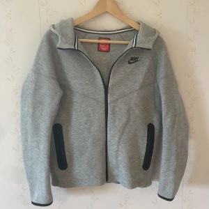 Nike tech Fleece grå - Snygg grå zip-hoodie från Nike med svart logga på bröstet och svarta dragkedjor vid fickorna. Jackan har huva, hel dragkedja och är långärmad. Perfekt för chill eller träning, riktigt clean look och skön passform. Ny pris 1200kr. Använt 1 gång.
