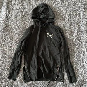 Svart hoodie från The Black Label Collection - Svart hoodie i 100% bomull från The Black Label Collection. Snyggt tryck med korsade nycklar på bröstet och texten 'KEYS TO FREEDOM' på ryggen. Huva med snörning, långa ärmar och dragkedjor i sidorna för extra edge.