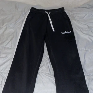 Svarta mjukisbyxor Black Squad XS - Svarta mjukisbyxor från Black Squad i storlek XS. Byxorna har vit snörning i midjan, en vit rand längs ena benet och texten 'Los Angeles' broderad på ena sidan. Perfekta för chill dagar eller när du vill ha en avslappnad look.
