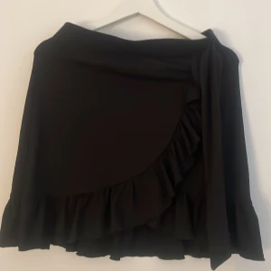 Svart omlottkjol med volang och knyt - Supersnygg svart kort omlottkjol med volangkant och knyt i sidan. Kjolen har en feminin och flowy känsla, perfekt att styla med en cool topp eller stickad tröja. Materialet känns lätt och följsamt.