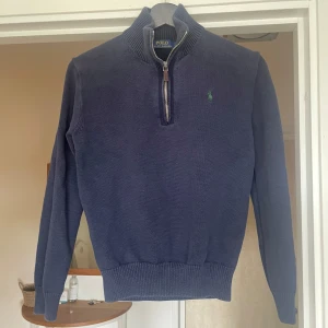 Marinblå stickad tröja Polo Ralph Lauren - Säljer min snygga marinblå stickade tröja från Polo Ralph Lauren med halv dragkedja och hög krage. Klassisk broderad logga i grönt på bröstet. Perfekt för lager-på-lager och passar till allt. Ribbstickade muddar och nederkant ger en clean look, samt väldigt bekväm. Passar en som är mellan 160-170. Hör av er vid intressen! 