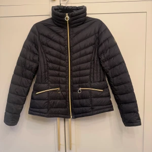 Svart dunjacka från Michael Kors - Lätt svart dunjacka från Michael Kors med guldfärgade dragkedjor och hög krage. Jackan har quiltad design, två fickor med dragkedja.