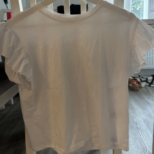 Vit volangärmad topp från Zara - Supersöt vit topp från Zara med volangdetaljer på ärmarna. T-shirten har rund halsringning och är tillverkad i mjuk bomull, perfekt för en fräsch och stilren look. Enkel att matcha med både jeans och kjol.