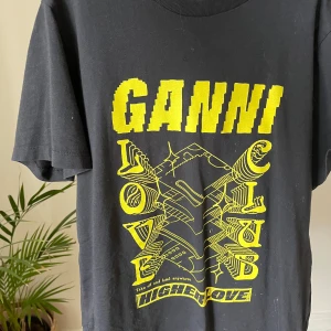 Svart Ganni t-shirt med gul print - Cool svart t-shirt från Ganni med stor gul grafisk print och texten 'HIGHER LOVE' framtill. Klassisk passform med rund hals och korta ärmar. Perfekt statement-plagg för dig som gillar streetstyle och unika tryck.