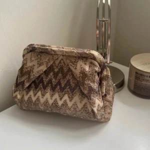 Beige kuvertväska från Beck Söndergaard - Säljer en kuvertväska från Beck Söndergaard i beige och bruna toner med snyggt zickzackmönster. Väskan är tillverkad i ett tygmaterial och har en klassisk form med metallspänne upptill. Perfekt att bära i handen för en cool och trendig look.