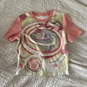 Färgglad kortärmad topp med konstmotiv - Unik topp med konstnärligt tryck i pastellfärger och motiv av en person i spiral. Ärmarna är ribbade och rosa, medan kroppen har ett färgstarkt mönster i rosa, grön, gul och lila. Rund halsringning och croppad passform🪅