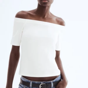 off shoulder topp - Säljer en stilren vit offshoulder topp med korta ärmar. Toppen har en rak och enkel passform som sitter snyggt över axlarna. Perfekt för dig som gillar minimalistisk och fräsch stil.
