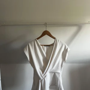 Vit omlottopp från Zara - Säljer en stilren vit omlottopp från Zara med knyt i midjan. Toppen har v-ringning och är ärmlös, vilket ger en fräsch och modern look. Perfekt att matcha med jeans eller kjol för en clean stil. Endast andvänd ett fåtal gånger
