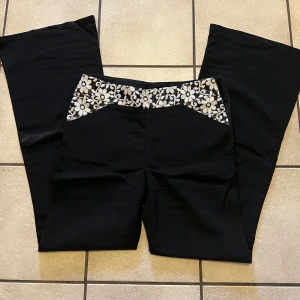 Svarta bootcut byxor med spetsdetalj - Svarta byxor med bootcut passform och snygg blommig spetsdetalj i midjan. Byxorna har en klassisk siluett med utsvängda ben och en unik touch med den vita spetsen upptill. Perfekta för dig som vill sticka ut lite extra. Midja omkrets 72 cm.