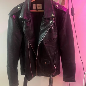 Svart skinnjacka från Deadwood, stl 44 - Riktigt snygg svart skinnjacka från Deadwood i klassisk bikerstil. Jackan har breda slag, dragkedjor, fickor med både dragkedja och knapp samt bälte i midjan. Perfekt för dig som vill ha en edgy och tidlös look. En mindre defekt på hängöglab som lossnat i en änden enkelt att laga om man vill.
