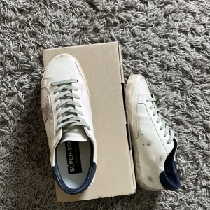 Golden Goose Superstar sneakers vit/grå - Golden Goose Superstar sneakers i vitt skinn med grå mockastjärna på sidan, grå snörning och mörkblå häl. Snygg vintage-look med slitna detaljer och bruna yttersulor. Perfekta för dig som gillar streetstyle och unika detaljer.