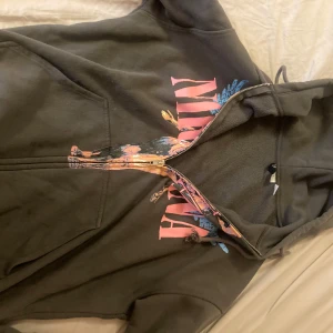 Svart Nirvana hoodie från H&M Divided - Svart hoodie med dragkedja från H&M Divided i samarbete med Nirvana. Stor tryckt grafik framtill med rosa text och motiv i blått och orange. Tröjan har huva med snörning och är långärmad. Perfekt för dig som gillar bandmerch och streetstyle.