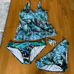 Blågrön bikini med tropiskt mönster - Bikini med tropiskt bladmönster i blått, grönt och svart. Setet består av en tankini-topp med snörning i sidan och två olika trosor, en med dekorativa band. 