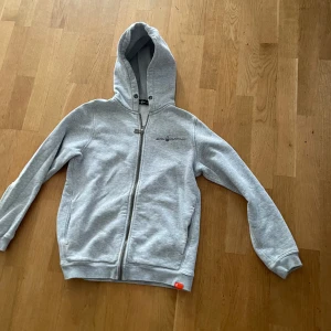 Grå hoodie från Sail Racing - Ljusgrå hoodie från Sail Racing med dragkedja framtill och huva. Tryck med logga på bröstet och stort tryck på ryggen. Mjuk bomullskänsla, ribbade muddar och fickor på sidorna. Perfekt för chill dagar eller när du vill ha en clean streetstyle.