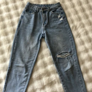 Ljusblå ripped jeans med hög midja - Säljer ett par ljusblå jeans med hög midja och raka ben. Jeansen har slitna detaljer vid fickan och ett större hål på ena knät för en cool, avslappnad look. Klassisk femficksmodell och bälteshällor. Perfekta till sneakers eller boots. Lös passform. Storlek M