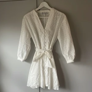Vit kortklänning från Zara - Superfin vit kortklänning från Zara med broderade blommor, knappar framtill och knytskärp i midjan. Klänningen har trekvartsärmar och v-ringning. Perfekt nu till sommaren💕