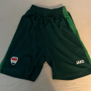 Gröna Jako fotbollsshorts Irak - Mörkgröna fotbollsshorts från Jako med ljusgröna sidor och Irak-emblem på ena benet. Elastisk midja och sportig passform, perfekta för träning eller match. Materialet är lätt och andas, vilket gör dem sköna att ha på sig under intensiva aktiviteter.