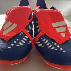 Adidas blå-röda fotbollsskor storlek 45 1/3 - använda en tränning. Snygga fotbollsskor från Adidas i blått och rött med vita detaljer och klassiska ränder. Skorna har snörning och täckande plös med Adidas-logga. Ovandelen är i syntetmaterial och sulan har dobbar för bra grepp på planen. Perfekta för dig som vill sticka ut på matchen.