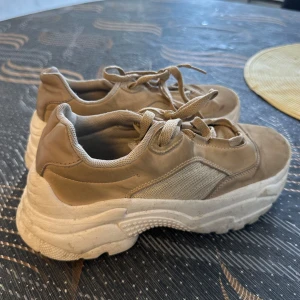 Chunky beige sneakers med plattform - Chunky sneakers i beige med plattformsula och snörning. Ovandelen är i en mix av mockaliknande material och mesh, vilket ger en cool kontrast. Snyggt sportig vibe och passar perfekt till streetwear. Tjock vit sula för extra attityd.