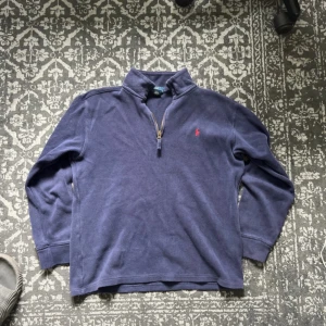 Blå half zip tröja från Polo Ralph Lauren - Mörkblå långärmad tröja från Polo Ralph Lauren med half zip-dragkedja och klassisk ståkrage. Broderad röd logga på bröstet. Tillverkad i mjuk bomull, perfekt för lager-på-lager under höst och vinter. Storlek L för pojkar (14-16 år)