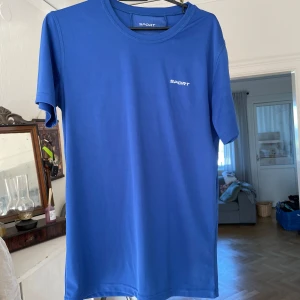 Träningströja - Blå t-shirt från Sport med kort ärm och rund hals. Tillverkad i ett lätt och stretchigt polyestermaterial, perfekt för träning. Diskret logga framtill och reflekterande detalj på ryggen. Enkel och clean design som passar dig som gillar sportiga vibes. Stlk S. Använd 1 gång!