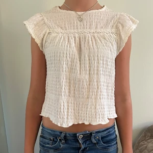 Vit blus med volangärm och spets - Vit blus i krinklad bomull med volangärmar och spetsdetalj över bröstet. Blusen är croppad och  har rund halsringning. Den är armlös med volangkanter.