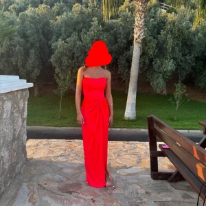 Röd/rosa axelbandslös långklänning - Säljer en röd/rosa axelbandslös långklänning med figurnära passform och snygg drapering i midjan. Klänningen har slits framtill och markerad byst, vilket ger en elegant och modern look. Perfekt för dig som vill sticka ut med en färgstark och stilren outfit. Du behöver heller inte oroar dig för att klänningen ska åka ner då denna är av hög kvalité och inte rörde sig hela kvällen. 