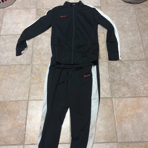 Svart Nike vindjacka med vita ränder - Svart vindjacka från Nike med vita ränder längs ärmarna och röd Nike-logga på bröstet. Jackan har dragkedja framtill och hög krage, perfekt för sportiga dagar eller när du vill ha en clean och sportig look.