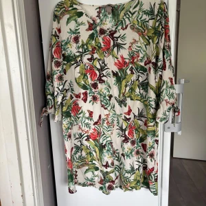 Blommig kortklänning med knytärm - Superfin blus med tropiskt blommönster i grönt, rött och vinrött på vit botten. Klänningen har trekvartsärmar med knyt i ärmslutet och en avslappnad passform. Perfekt för sommarens alla tillfällen när du vill sticka ut lite extra. 