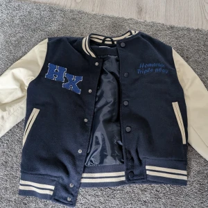 Blå och beige bomberjacka HX - Snygg blå och beige bomberjacka från HX med broderad text och stora bokstäver på bröstet. Jackan har kontrastärmar, randiga muddar och knappar framtill. Perfekt för en sportig och trendig look.