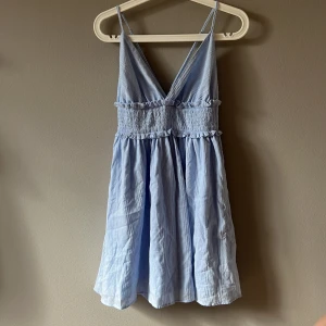 Ljusblå kort sommarklänning - Supersöt ljusblå kortklänning med smala axelband och v-ringning både fram och bak. Klänningen har smockad midja, volangdetaljer och är tillverkad i ett lätt, luftigt bomullstyg. Perfekt för varma dagar och ger en fräsch vibe.