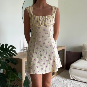 Ljusgul blommig klänning  - Supersöt kortklänning i ljusgul med små  blommor. Klänningen har breda axelband som man kan ha antingen uppe på axlarna eller ” dra ner” på sidan av axlarna/armarna.