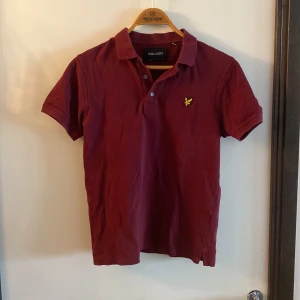 Vinröd pikétröja från Lyle & Scott - Snygg vinröd pikétröja från Lyle & Scott med klassisk krage och knappslå. Tröjan har korta ärmar och den ikoniska gula loggan broderad på bröstet. Perfekt för en stilren och avslappnad look.