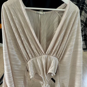 Plisserad champagnefärgad body NLY Trend - Säljer en plisserad body från NLY Trend i en lyxig champagnefärg. Toppen har djup v-ringning, vida ärmar och ett snyggt omlottliknande fall framtill. Materialet är glansigt och ger en cool, trendig vibe. Perfekt för dig som vill sticka ut lite extra!