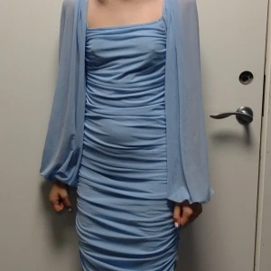 Ljusblå sexig omlottklänning med puffärm - Supersnygg ljusblå kortklänning med draperad effekt och transparenta puffärmar. Klänningen har fyrkantig urringning och sitter tight över kroppen. Perfekt för dig som vill sticka ut med en trendig look. Materialet känns lätt och luftigt.