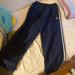 Mörkblå Adidas träningsbyxor med ränder - Säljer ett par mörkblå träningsbyxor från Adidas med klassiska vita ränder längs sidorna och broderad logga på låret. Byxorna har elastisk midja och är tillverkade i ett lätt och sportigt material, perfekta för chill eller träning.