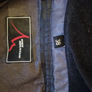 Lila vindjacka från High Mountain - Snygg lila vindjacka från High Mountain med huva och flera dragkedjeförsedda fickor. Jackan har en sportig look och är perfekt för utomhusaktiviteter. Långärmad modell med coola detaljer och kontrastfärgade dragkedjor.