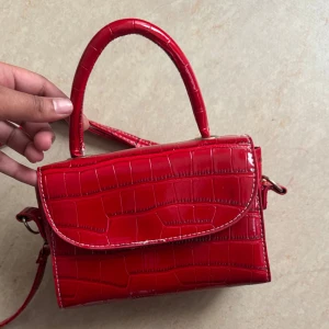 Röd croco handväska med axelrem - Snygg röd handväska med croco-mönster och glansig finish. Väskan har både handtag och avtagbar axelrem, perfekt för att bära på olika sätt. Stilren och trendig design som lyfter vilken outfit som helst.