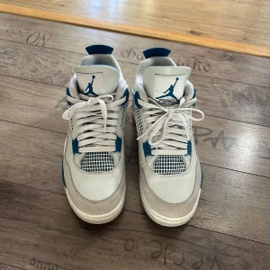 Nike Air Jordan 4 Retro Vit/Blå - Säljer ett par Nike Air Jordan 4 Retro i vitt och blått med storlek 42,5. Klassisk siluett med meshpaneler, snörning och detaljer i blått på häl, sula och snörning. Ovandel i skinn och syntet, med ikonisk Jumpman-logga på plösen och Nike Air på hälen. Perfekta för dig som älskar sneakers med retro vibe.