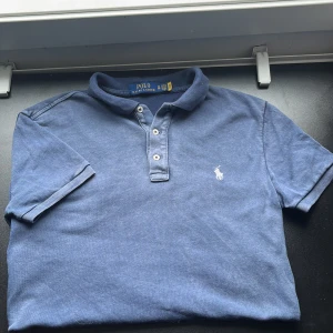 Blå pikétröja från Polo Ralph Lauren XS - Klassisk blå pikétröja från Polo Ralph Lauren i storlek XS. Tröjan har korta ärmar, krage och tre knappar framtill. Ikoniska broderade loggan i vitt på bröstet. Ser väldigt urtvättad ut men det är ljuset:) använd fåtal gånger och den är tillverkad i mjuk bommul. Undrar ni något så hör av er!👏