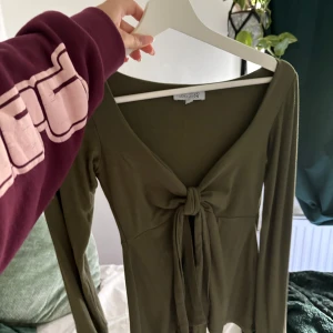 Olivgrön långärmad knytblus - Supersnygg olivgrön långärmad topp från design by si med djup v-ringning och knyt framtill. Tunn och mjuk i materialet, perfekt till jeans eller kjol. Sitter figurnära och har en modern, enkel look.
